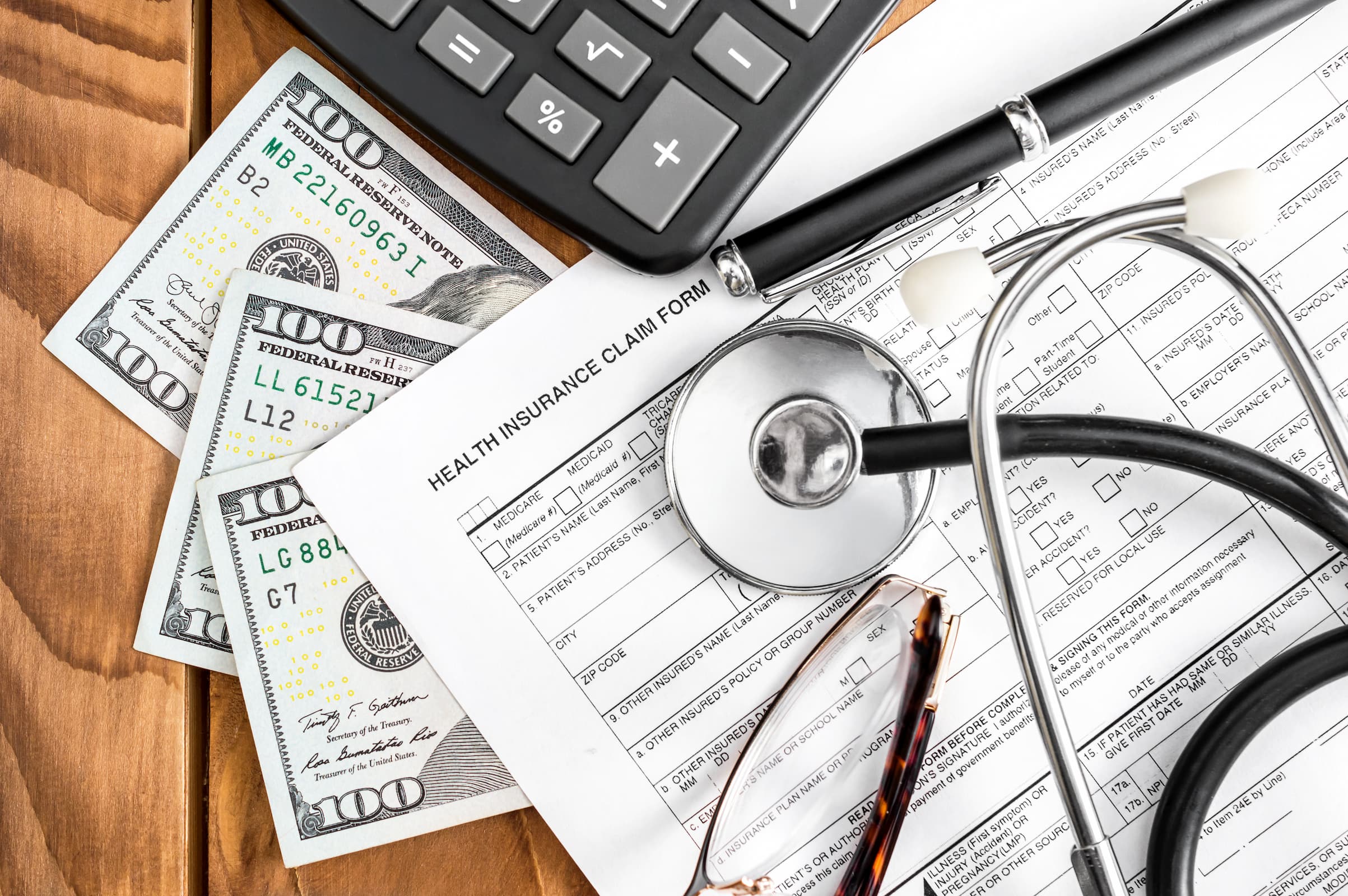 A health insurance claim form rests on a wooden surface, accompanied by a stethoscope, eyeglasses, a calculator, and several U.S. 100-dollar bills—items often found in the realm of personal injury lawyers navigating complex claims.