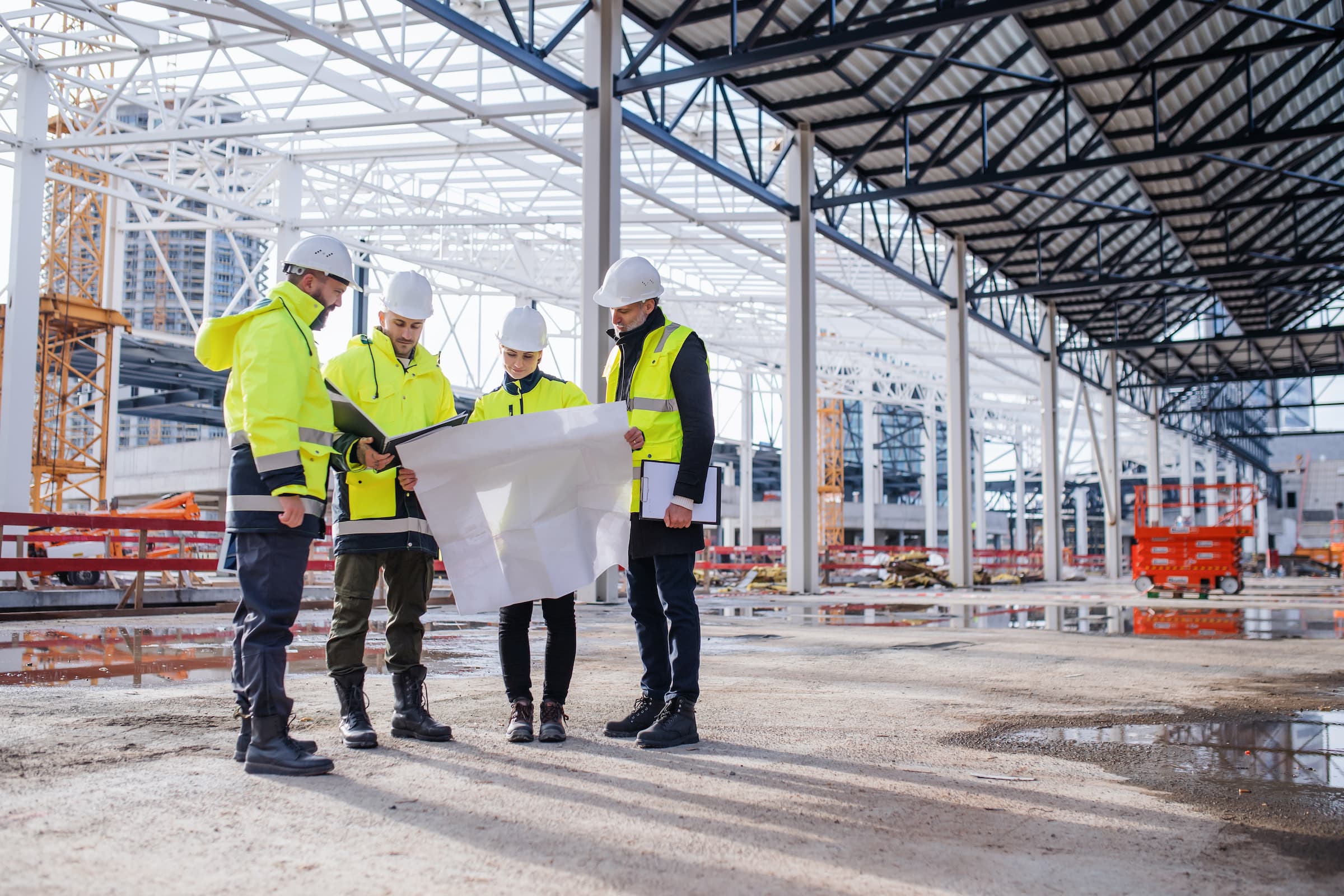 Four construction workers in high-visibility jackets and hard hats stand under a large metal structure, examining a blueprint. With construction equipment in the background, this partially built facility highlights the importance of safety—a concern often addressed by construction accident attorneys.