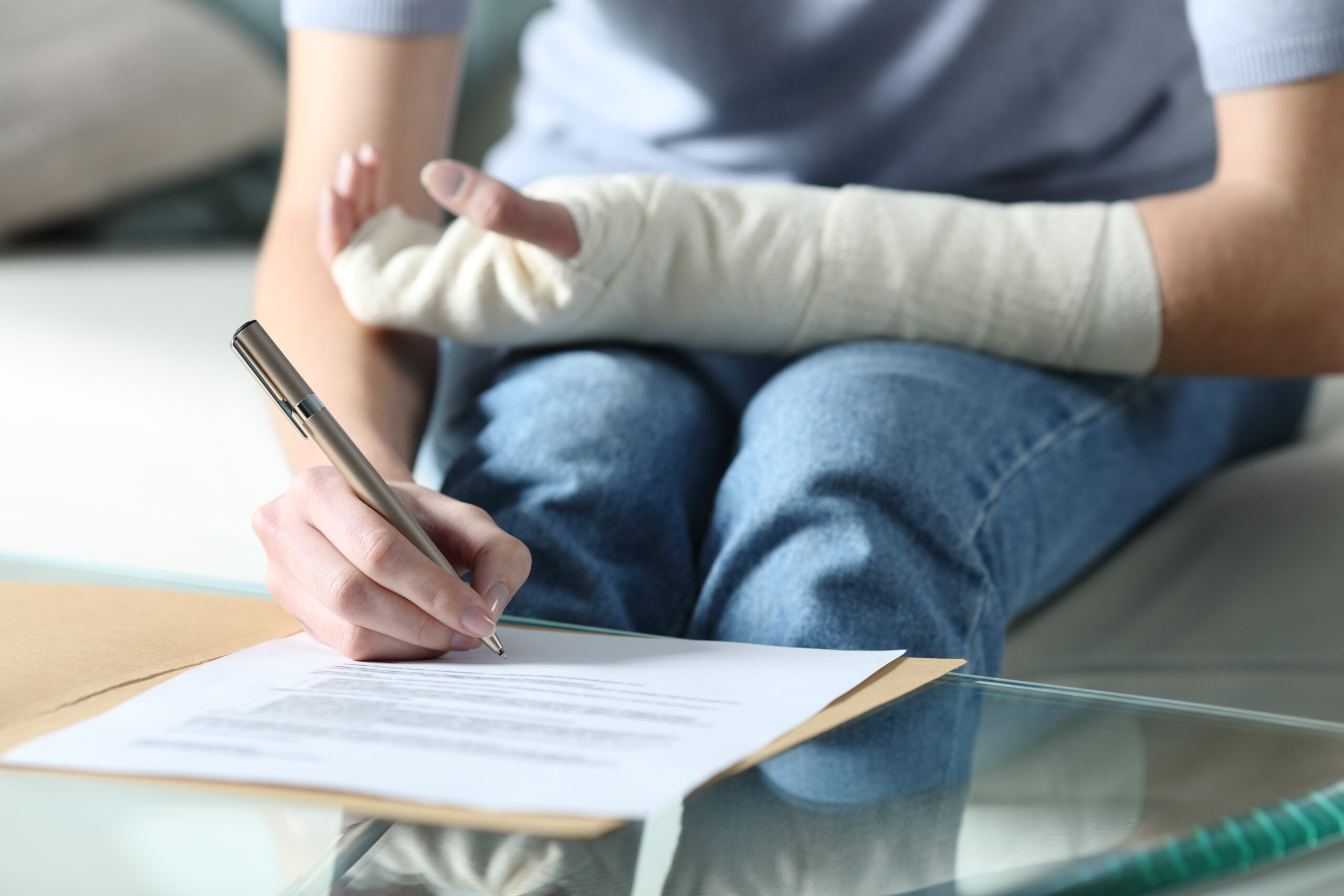 A person with a cast on their right arm is sitting on a couch, using their left hand to write on a piece of paper placed on a glass table. They are wearing a light blue shirt and jeans. An envelope, possibly related to personal injury claims, is also visible on the table.