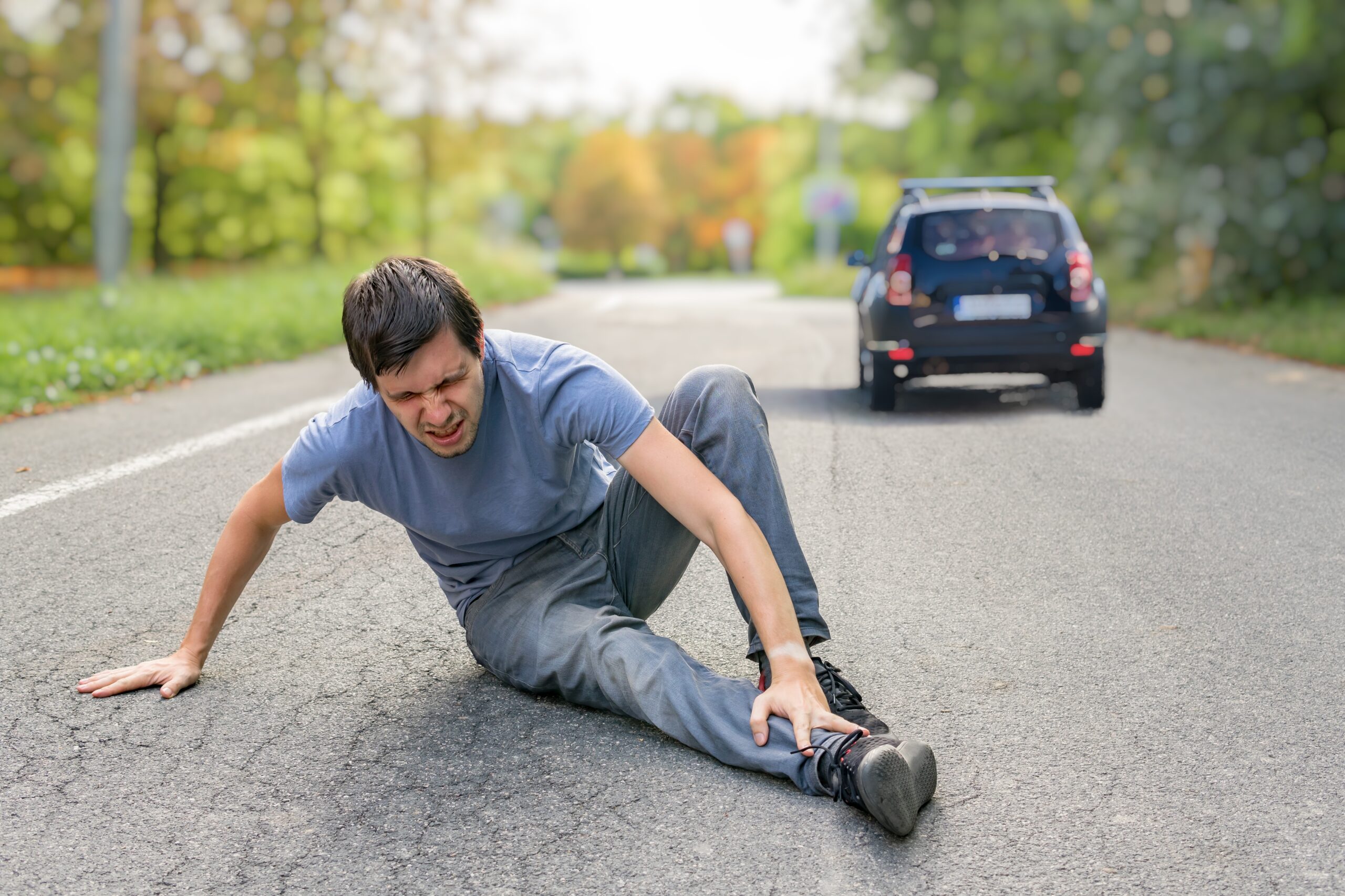 A man in a gray t-shirt and pants is sitting on the ground in the middle of a road, clutching his ankle with a pained expression. A small black car, seemingly involved in an auto draft situation, is seen driving away from the scene. The road is surrounded by greenery.