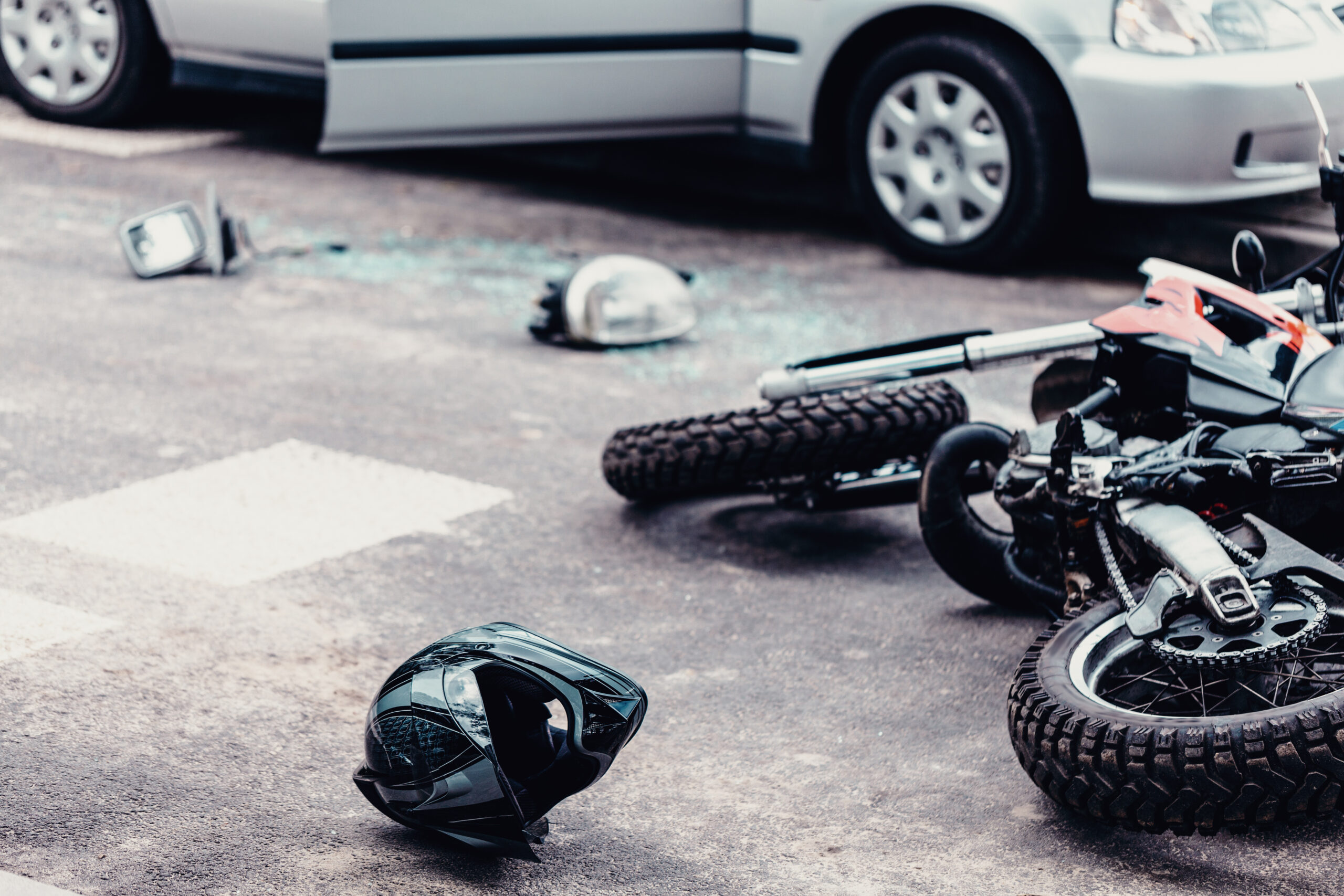 A motorcycle lies on its side next to a broken car mirror and a helmet on the pavement. A silver car with visible damage is parked nearby. Shattered glass and debris are scattered on the ground, highlighting the unique risks associated with motorcycle accidents.